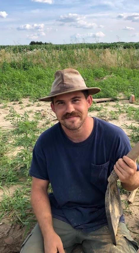 Picture of a seated man in a hat at an archaeological site, with long grass and a light blue cloudy sky behind him